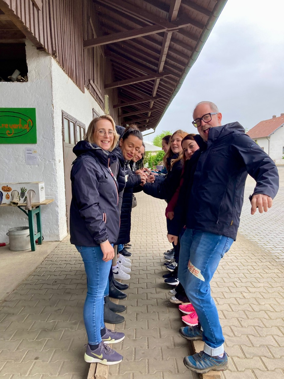 Chef und Mitarbeiterinnen stehen gemeinsam am Holzbalken- geben sich Halt und Sicherheit bei dem Geschicklichkeitsspiel