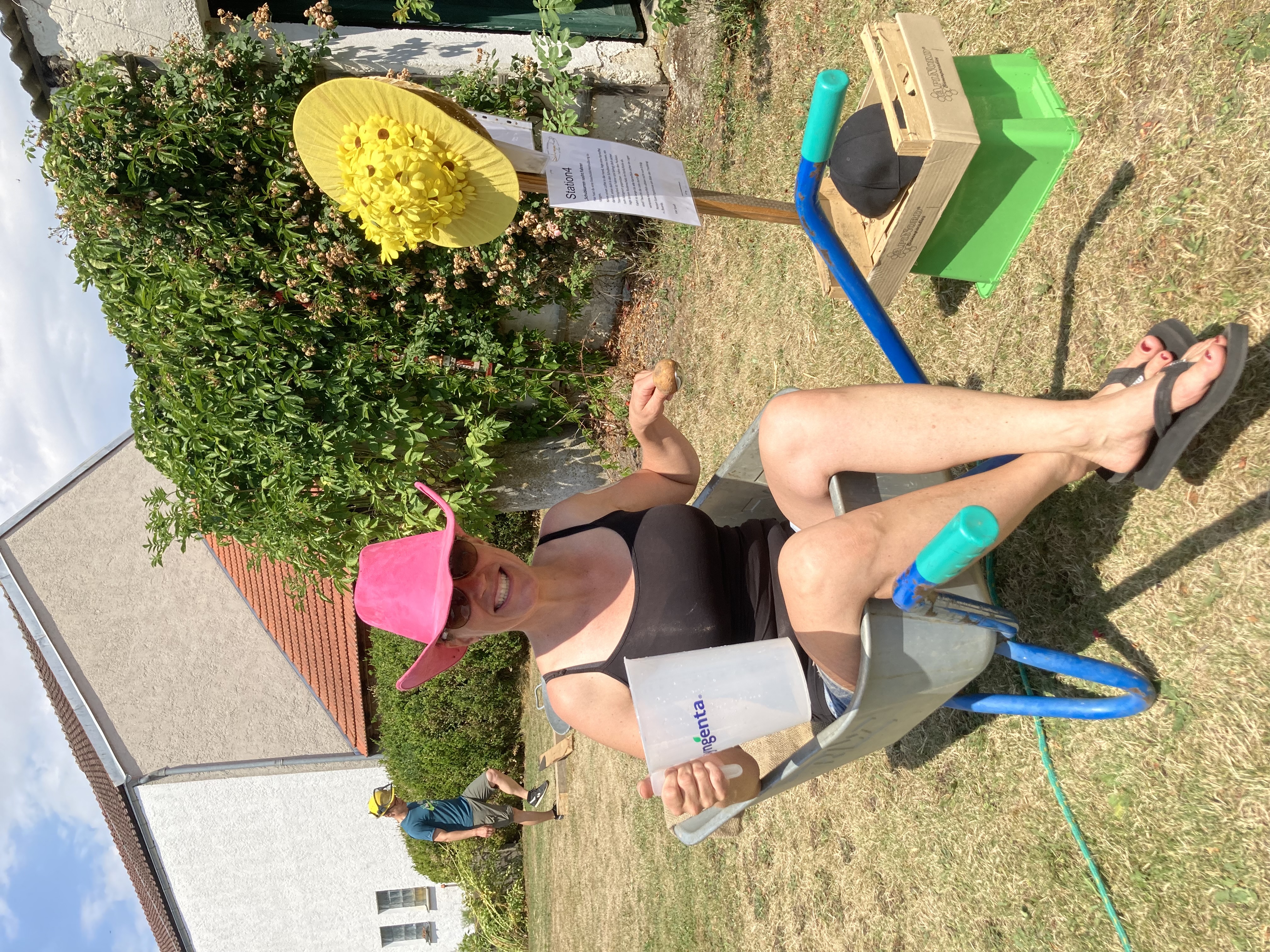 teilnehmerin-sitzt-im-schubkarren-hält-gegenstand-in-der-hand-firmenevent-bauernhofolympiade-idealer-ort-fuer-teamevent-nahe-straubing-fuer-grosse-gruppen-ideal-fahrt-am-pacour